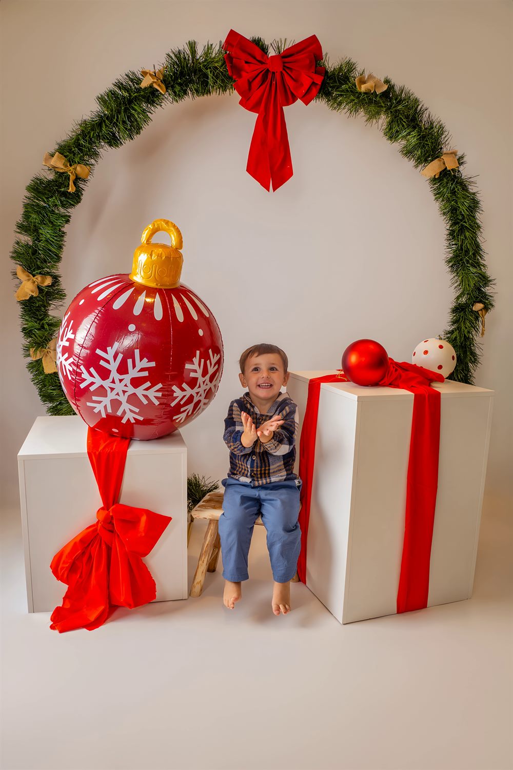 Niño aplaudiendo entre regalos gigantes y una bola roja bajo la corona navideña
